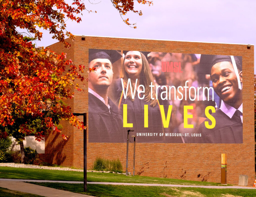 Large banner on a brick building reads "We transform lives" with photos of three graduates; University of Missouri-St. Louis campus with autumn leaves in foreground.