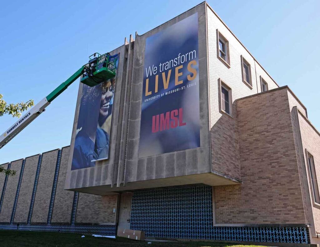A worker in a lift installs a large banner reading "We transform lives. University of Missouri–St. Louis. UMSL" on the exterior of a brick building.
